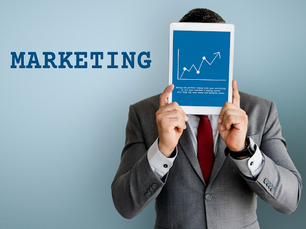 A man in a suit holds a tablet with a growth chart over his face. The word “MARKETING” appears on the wall to the left in bold blue text against a plain background.