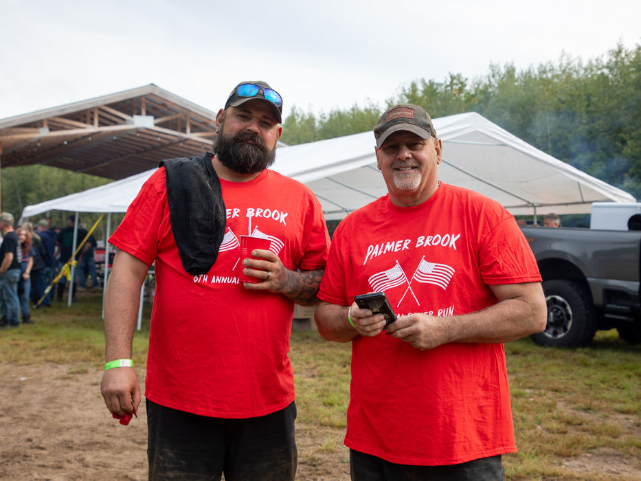 Two men wearing matching red shirts, holding beverages and smiling at the camera person.