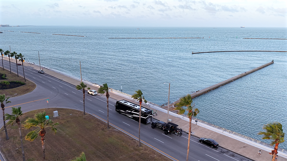 Coastal road with RV, trucks, and cars under palm trees. Ocean piers extend into calm blue water. Cloudy sky, peaceful scene.