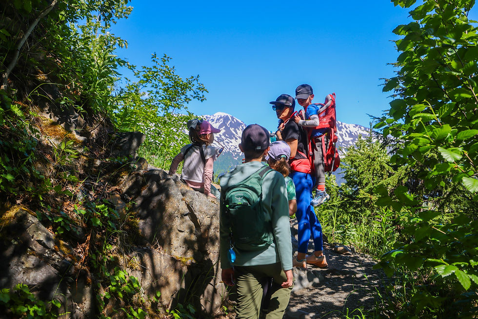 Family hikers with children on a rocky forest trail, one child in a red Deuter Kid Comfort Kid carrier, a boy with an Osprey Raptor day bag with snowy mountains under a bright blue sky.