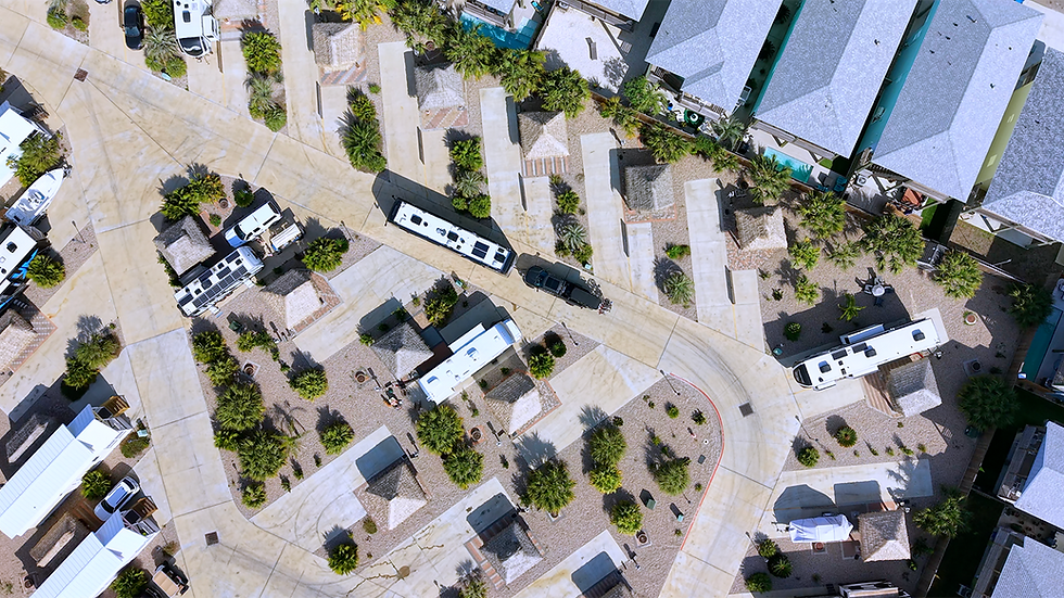 Aerial view of an RV park with organized rows of RVs and palm trees. Beige roads intersect among green patches and small cabins.