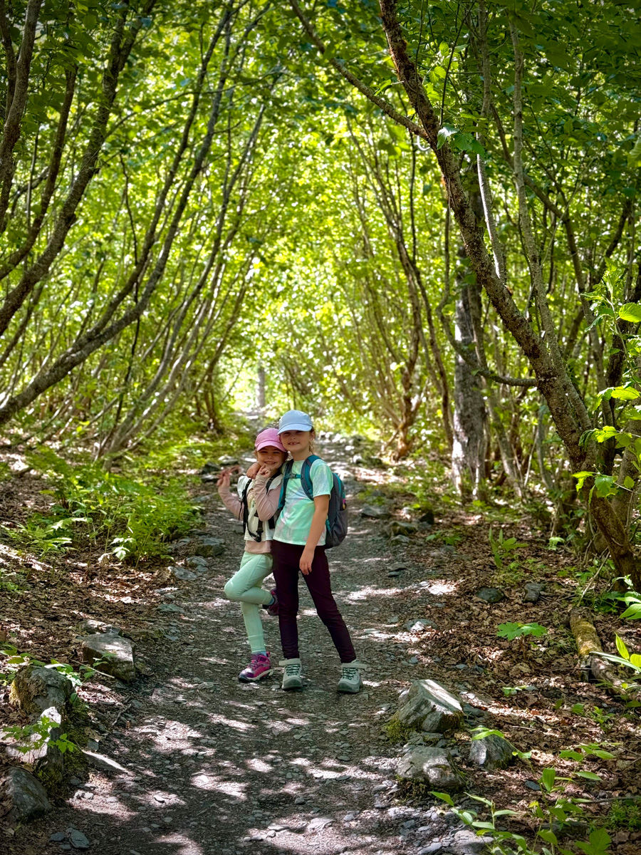 Two young girls with backpacks pose on a sunny forest trail under a lush green tree canopy.