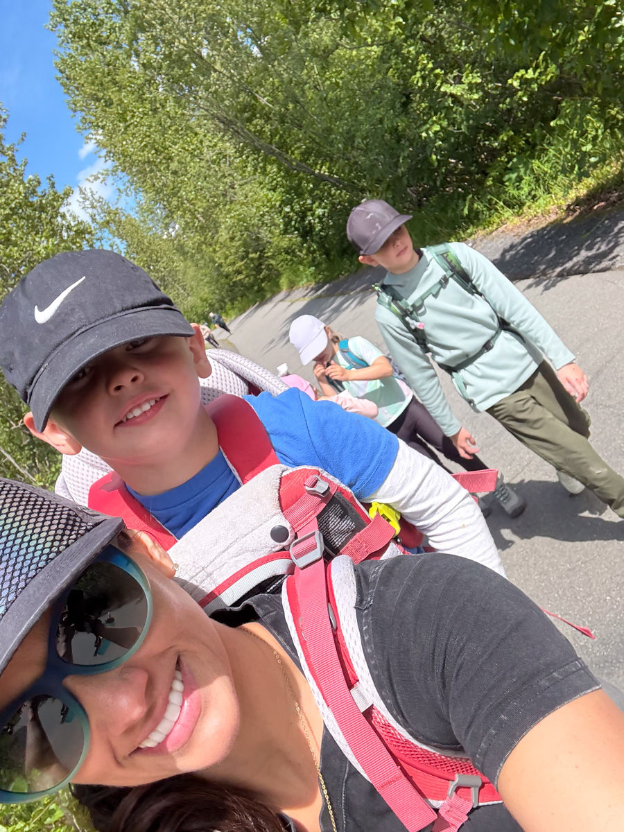 Smiling woman and children hiking on a sunny tree-lined path, wearing caps, backpacks, and carrying gear.