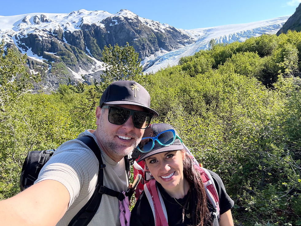 Smiling hikers take a selfie in front of snowcapped mountains and exit glacier under a bright blue sky on the Harding Icefield Trail in Kenai Fjords National Park.