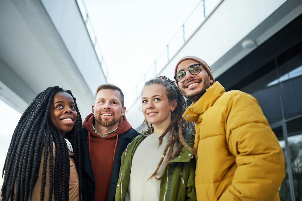 Four friends in winter clothes smile under a bridge. The bright sky, gray structure, and colorful jackets create a cheerful mood.