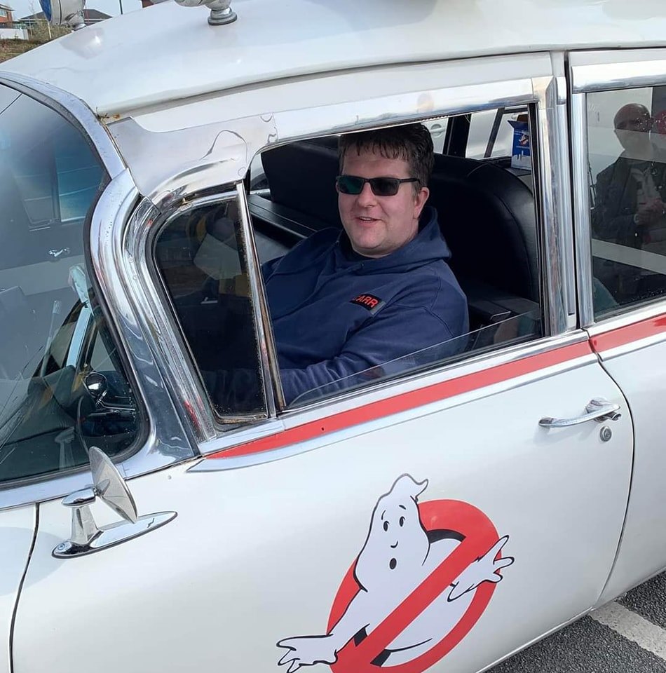 Young man in Ghostbusters car with logo and text smiling.