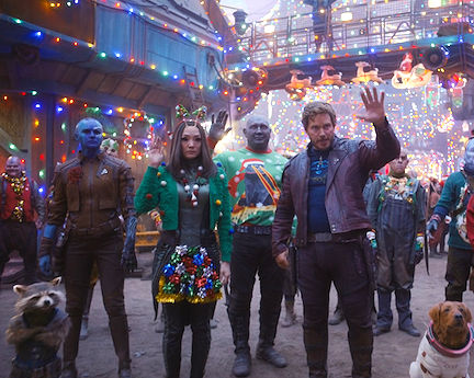Guardians of the Galaxy characters Nebula, Rocket Raccoon, Mantis, Drax, Peter Quill, and Cosmo the Spacedog all dressed in holiday festive attire standing in front of a decorated holiday lights while waving