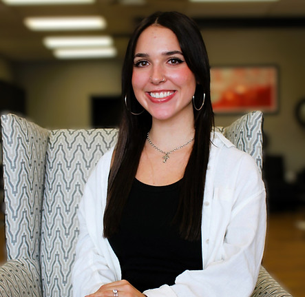 Smiling young woman seated in an armchair, wearing black top and a white jacket.