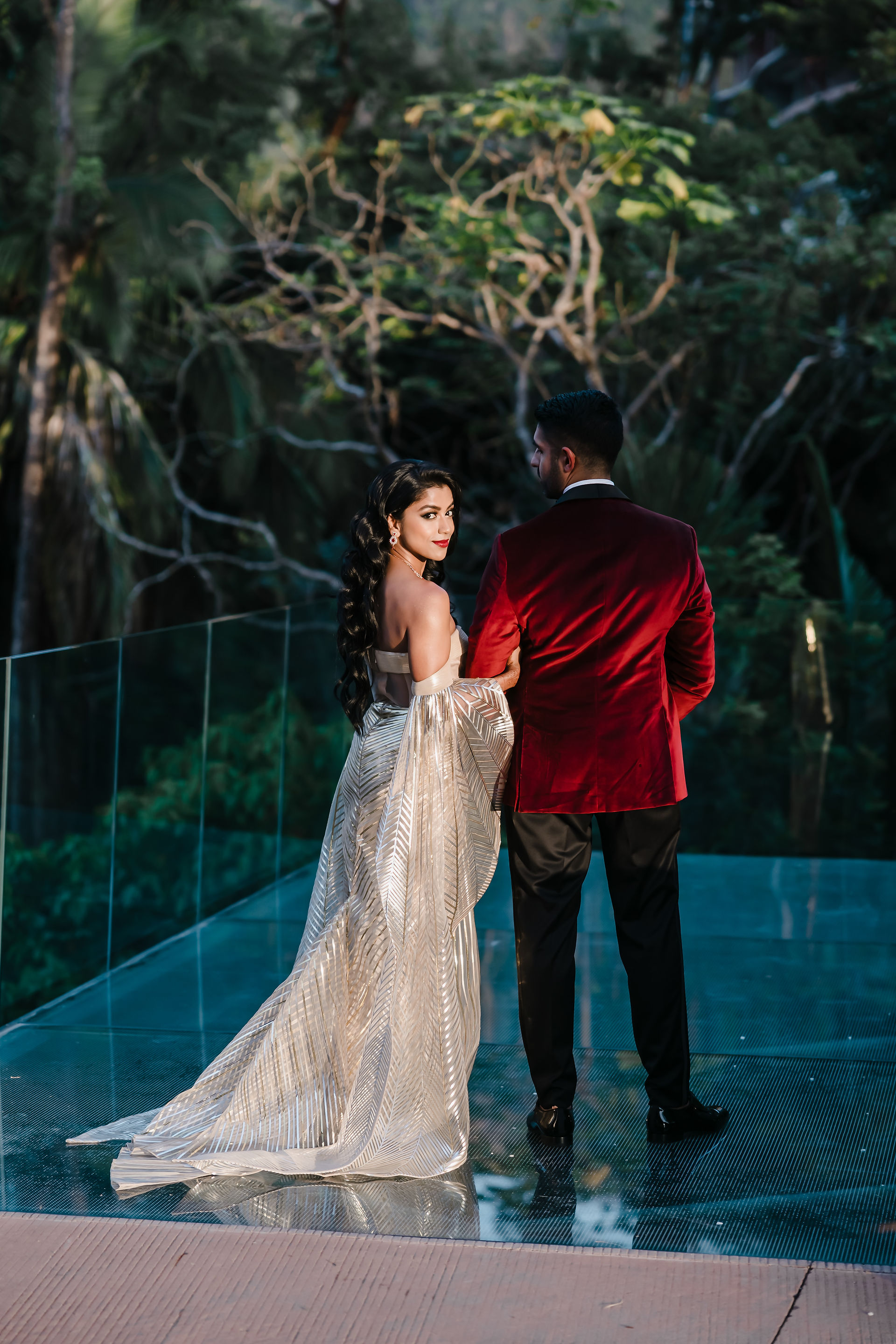 Stylish reception portrait of Bushra and Mohamed at Garza Blanca Puerto Vallarta. The bride, in a shimmering metallic gold gown with a dramatic patterned cape train, walks arm in arm with the groom, who wears a deep red velvet tuxedo jacket. She looks back over her shoulder with a confident smile, her long waves cascading down her back, while lush greenery frames the scene.