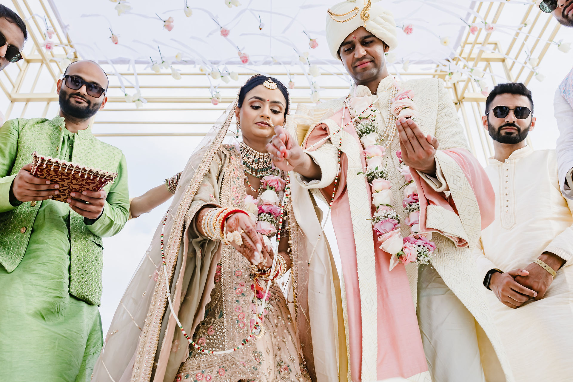 Devina and Rishabh during their Indian wedding ceremony at Moon Palace Cancún, holding hands as they perform a sacred ritual under a mandap adorned with hanging flowers, symbolizing blessings and union.