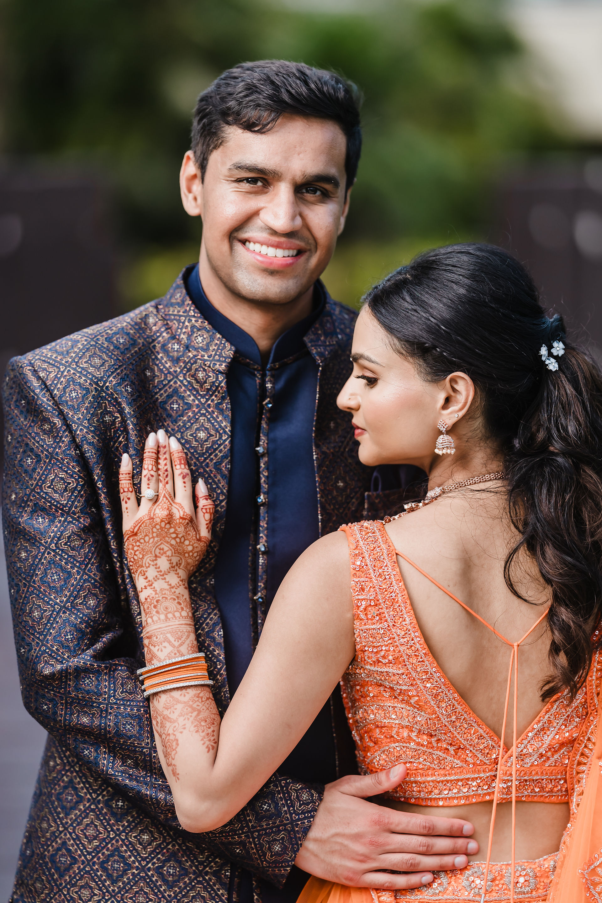 Rishabh smiles warmly in a black patterned sherwani as Devina, in her orange embroidered lehenga and diamond jewelry, leans into him with her mehndi-decorated hand resting on his chest during their Sangeet at Moon Palace Cancún.