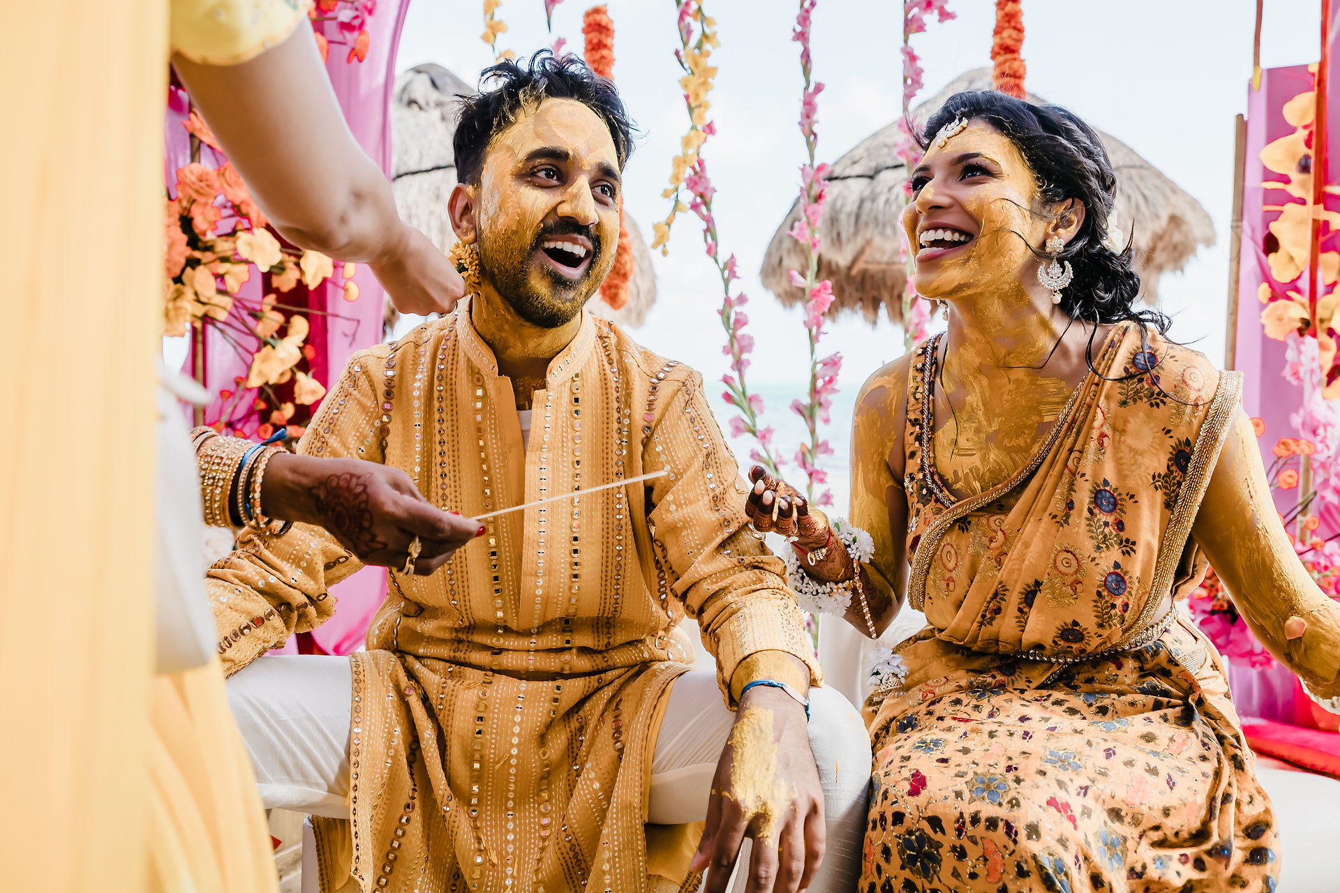 Bride and groom laughing together during their Haldi ceremony at Garza Blanca Cancun, both covered in golden turmeric paste under a colorful floral mandap.