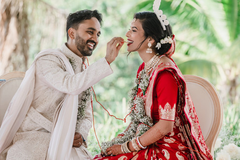 Groom feeds his bride during their Bengali Hindu wedding ceremony at Garza Blanca Cancun. He wears an ivory embroidered sherwani, and she is dressed in a vibrant red saree with gold embroidery, adorned with floral garlands, bridal jewelry, and a traditional mukut headpiece, both smiling joyfully against a tropical palm backdrop