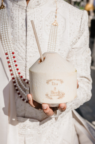 Groom in an embroidered white sherwani holding a personalized coconut drink engraved with “Rhia & Bhavik 12/14/2024” during their Garza Blanca Cancun destination wedding.