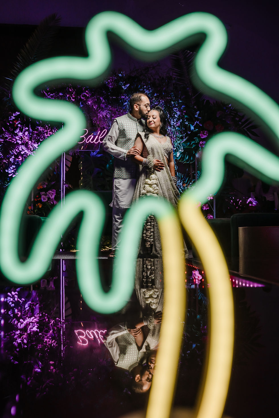 Romantic moment between bride and groom during their Indian destination wedding in Mexico, framed creatively through the glowing outline of a neon palm tree. The couple stands on a reflective black dance floor, their elegant silver and sage green outfits shimmering under vibrant purple and pink uplighting. Behind them, lush tropical florals and a neon sign add a festive, modern touch, while their mirrored reflection beneath enhances the dreamy, celebratory atmosphere of the night.