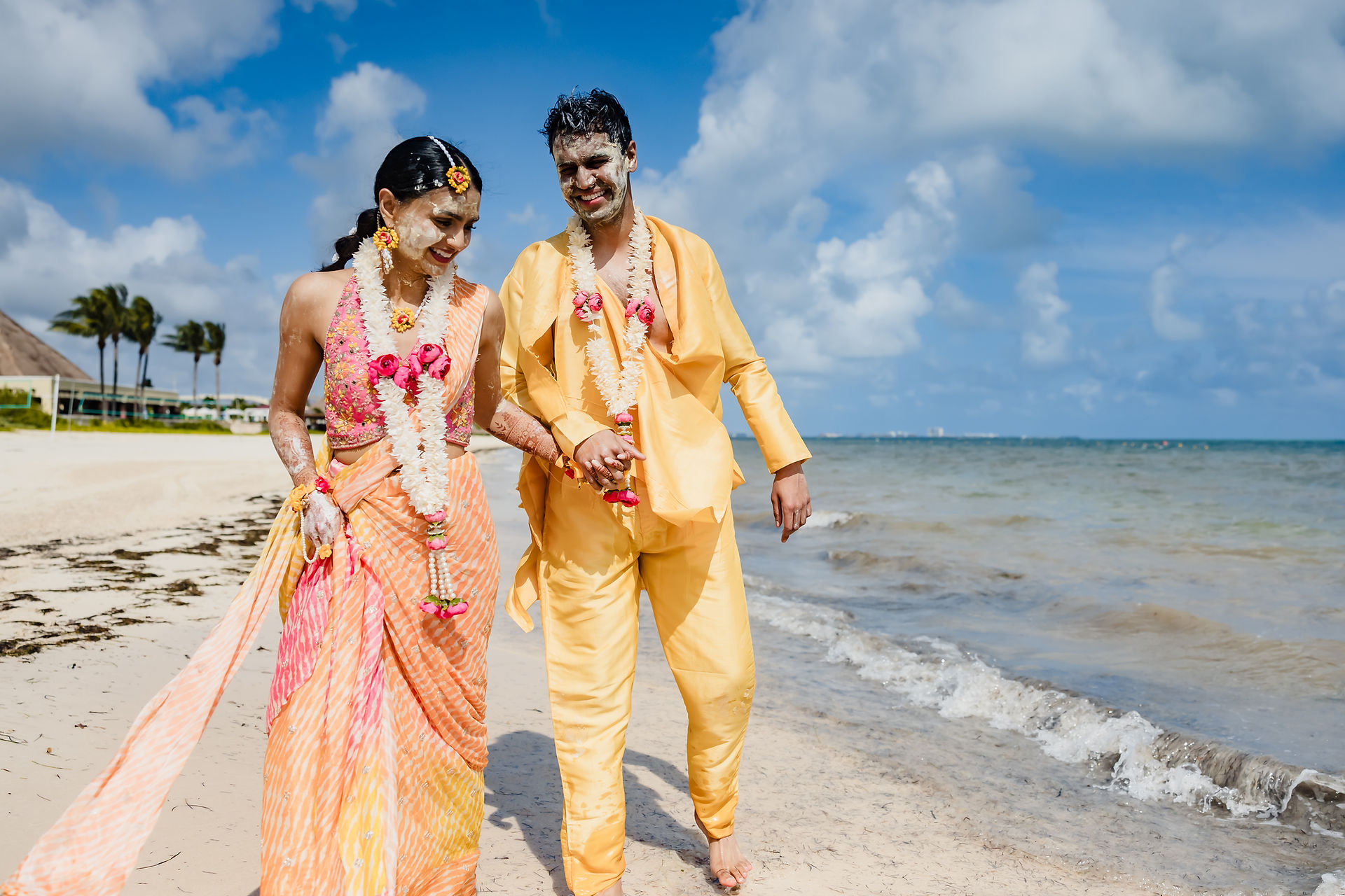 Devina and Rishabh, smiling and covered in haldi, walk hand in hand along the sandy beach at Moon Palace Cancún, dressed in peach and yellow outfits with floral garlands, the turquoise ocean and blue sky behind them.