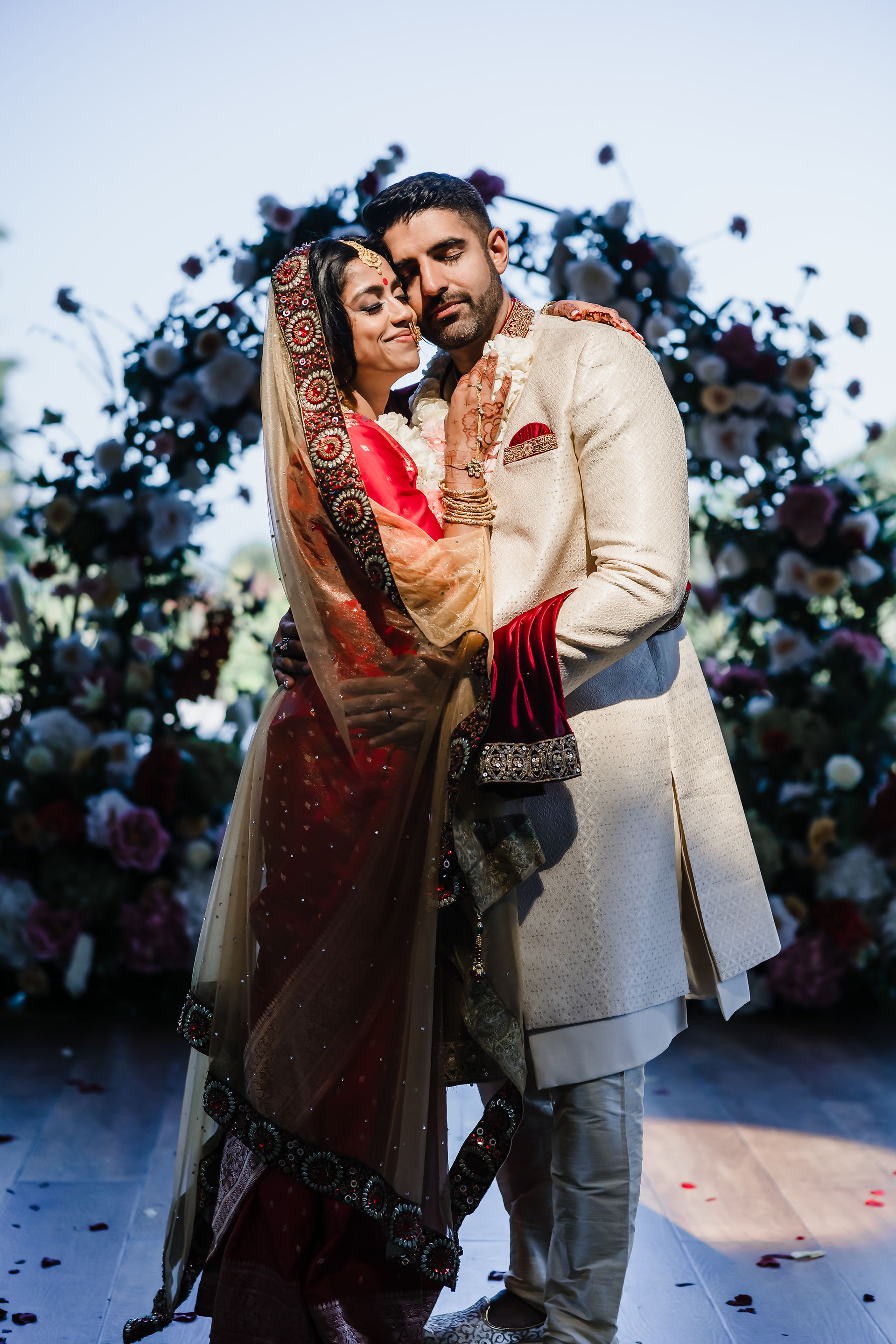 Romantic close-up of Bushra and Mohamed during their Garza Blanca Puerto Vallarta wedding. The bride, in a rich red saree with intricate gold embroidery and a beaded veil, rests her forehead gently against the groom’s temple. The groom wears an ivory sherwani with deep red velvet accents. Soft sunlight highlights their embrace, with a blurred floral arch of roses and peonies in the background, capturing pure love and serenity.