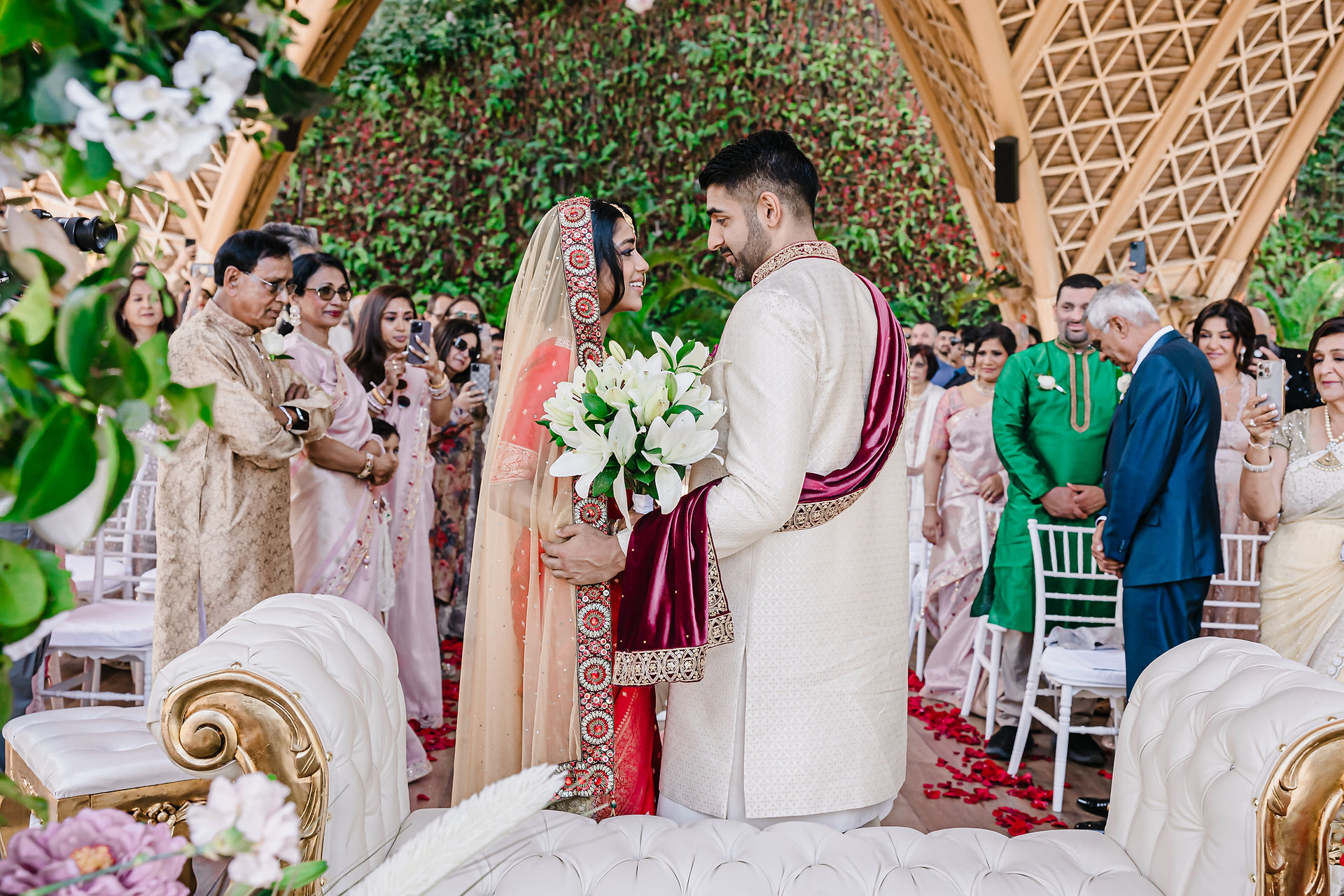 Bride in a traditional red and gold Bengali saree walks down the aisle at Tierra Luna, Garza Blanca Puerto Vallarta, holding a bouquet of white lilies. She smiles warmly at the groom, who is dressed in an ivory sherwani with a deep maroon stole, as they meet at the altar. Guests in elegant pastel and jewel-toned attire watch with joy, framed by lush tropical greenery and a bamboo-arched mandap adorned with flowers.