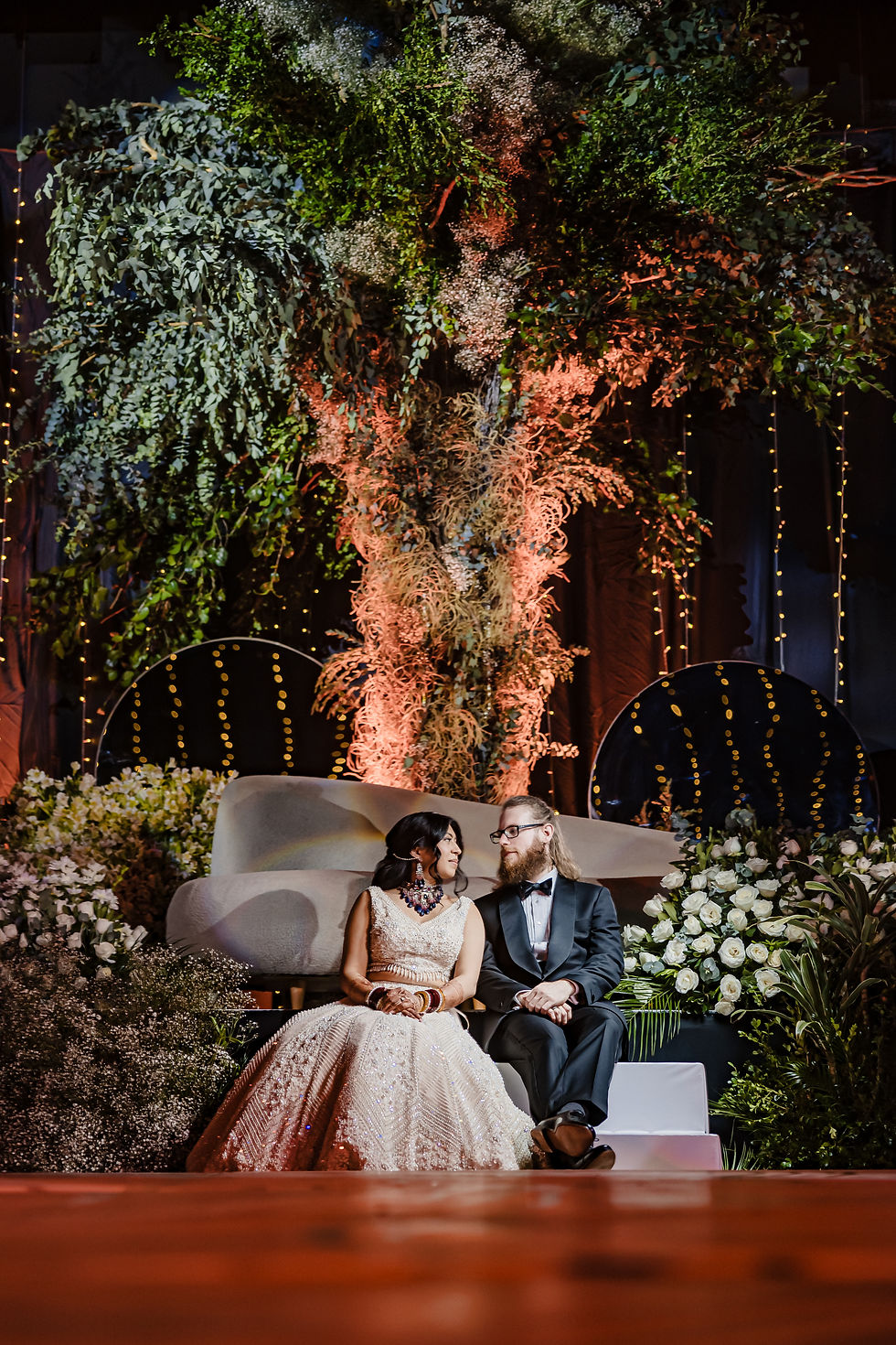 Bride and groom seated under a dramatic floral installation at their Indian wedding reception at Dreams Bahia Mita