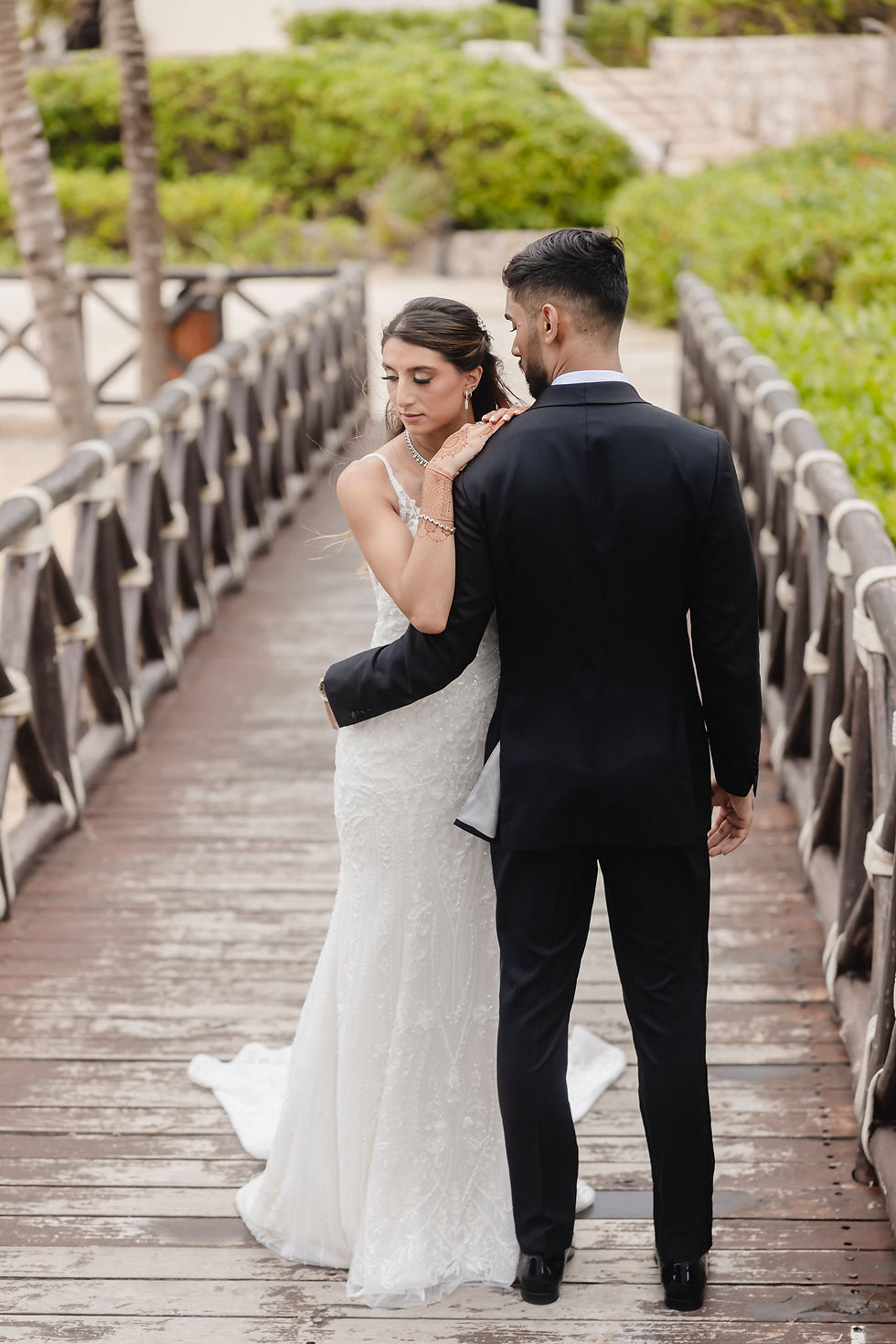 Bride in elegant white gown embraces groom in black tuxedo on wooden bridge at beachside wedding in Mexico.