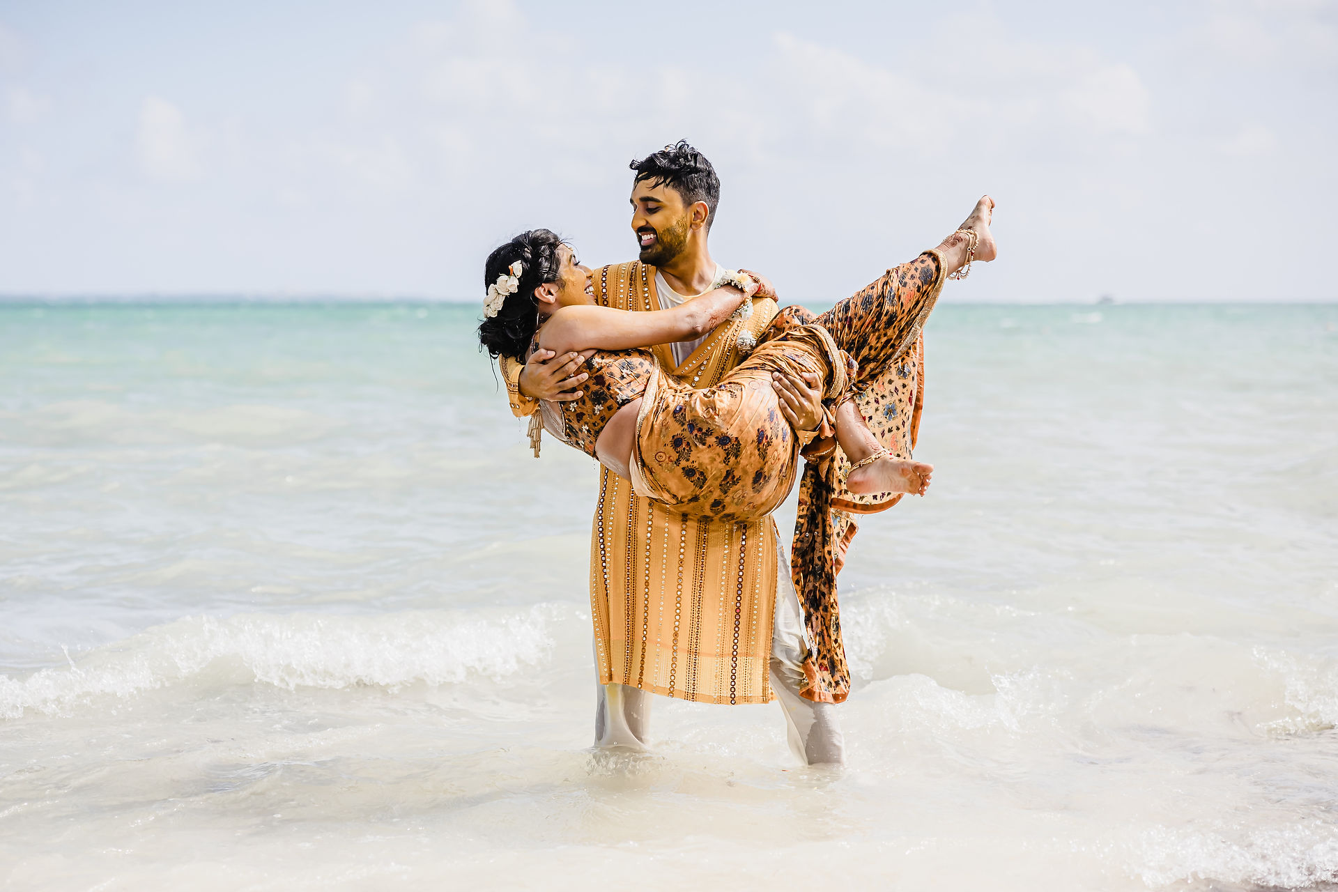 Groom, covered in turmeric from the Haldi ceremony, carries his smiling bride in the ocean at Garza Blanca Cancun, both wearing coordinated golden outfits.