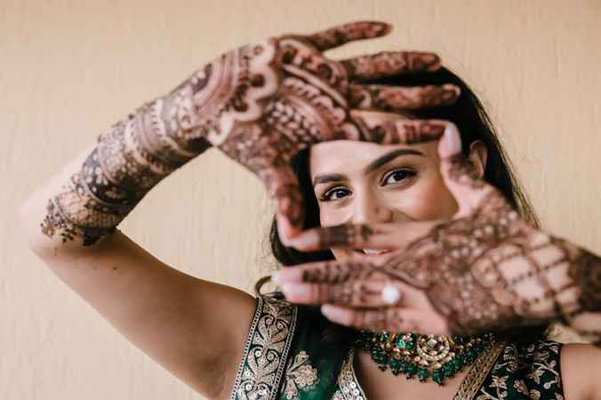 Bride Devina in an emerald green lehenga at Moon Palace Cancún playfully frames her face with her arms, showing intricate mehndi designs that stretch from her fingertips to her elbows. She smiles with joy, capturing the spirit of her Mehndi celebration.