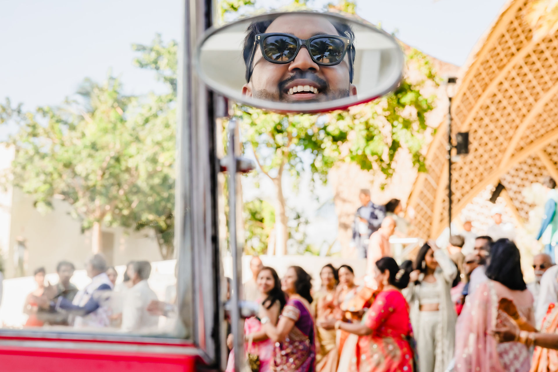 Reflection of the groom smiling in a vintage car mirror during his baraat at Garza Blanca Cancun, surrounded by guests in colorful Indian wedding attire.