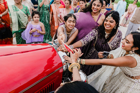 Guests and bridesmaids laugh and reach out to Devina as she departs in a vintage red car during the vidaai ceremony at Moon Palace Cancún, capturing a mix of tears, laughter, and celebration.