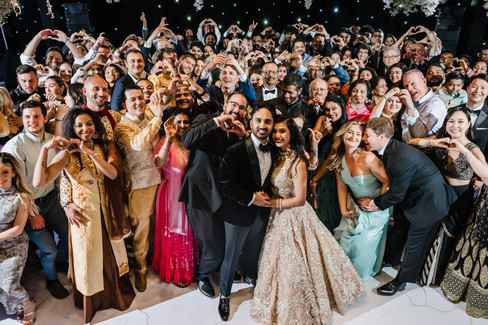 Bride and groom pose at the center of a joyful group photo during their Garza Blanca Cancun wedding reception, surrounded by guests making heart shapes with their hands, celebrating love and unity.