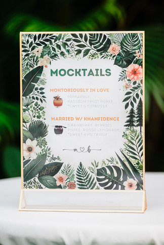 Tropical-themed mocktail menu sign at Bushra and Mohamed’s Garza Blanca Puerto Vallarta wedding, framed with lush green leaves and pink flowers. Features signature drinks named “Mohtoriously in Love” with pineapple and passion fruit puree, and “Married w/ Khanfidence” with cranberry, berry puree, and rose lemonade.