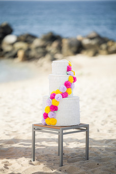 Three-tier wedding cake at Garza Blanca Puerto Vallarta, designed with white textured layers and adorned with a cascade of bright pink, yellow, and lavender flowers, set on the sandy beach with the ocean in the background.