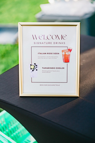 Framed welcome sign at Devina and Rishabh’s Moon Palace Cancún welcome party displaying their custom signature drinks. The menu includes “Italian Rose Soda” garnished with rose petals and “Tamarindo Cooler” inspired by tropical flavors. The chic white and gold frame sits on a black cocktail table with greenery in the background, adding a personalized touch to the celebration.
