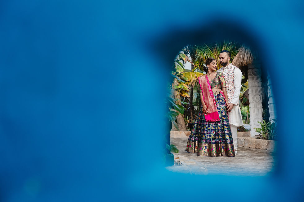 Creative portrait of an Indian couple in vibrant outfits at El Dorado Royale, framed through a keyhole-shaped cutout with tropical foliage and architectural elements in the background.