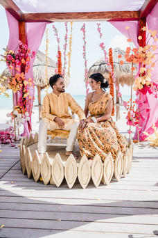 Indian bride and groom share a joyful moment during their Haldi ceremony at Garza Blanca Cancun, seated under a vibrant pink and orange floral mandap by the beach.
