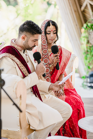 Bushra and Mohammed seated closely together during their Nikkah ceremony at Garza Blanca Puerto Vallarta, each holding a microphone as the groom reads aloud from a folded sheet of paper. The groom wears an ivory sherwani with a maroon velvet stole embroidered in gold, while the bride, dressed in a vibrant red saree with intricate gold embroidery, listens attentively under the airy bamboo arches of Tierra Luna.