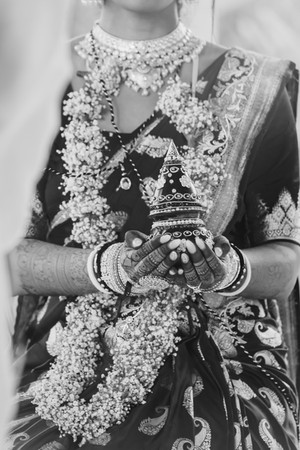 Close-up of an Indian bride in a red and gold saree holding a decorated kalash during her Hindu wedding ceremony at Garza Blanca Cancun, Mexico. Her hands are adorned with intricate mehndi designs, bangles, and jewelry, with fresh floral garlands draped around her neck, capturing a meaningful moment of Indian wedding traditions in a luxury destination setting.