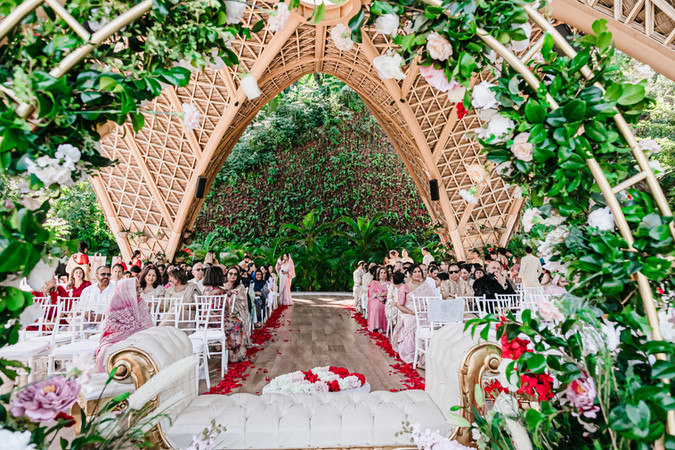 Wide-angle view of guests seated for a destination wedding ceremony at Tierra Luna, Garza Blanca Puerto Vallarta. The aisle is lined with red rose petals, leading to a lush green backdrop framed by a grand bamboo arch decorated with white and blush flowers. The scene captures the vibrant, tropical elegance and joyful anticipation of the celebration.