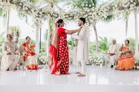 Bride in a red and gold Bengali saree placing a floral garland around the groom’s neck during their wedding ceremony at Garza Blanca Cancun, surrounded by white flowers and palm trees.