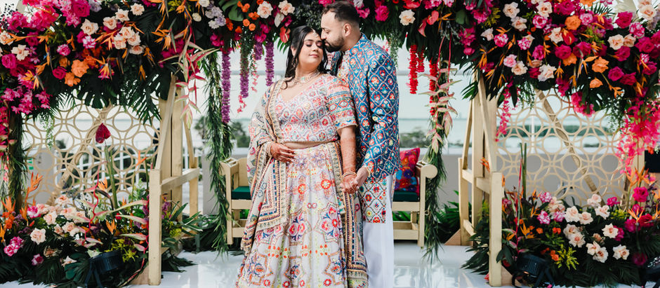 Indian couple posing beneath a vibrant floral arch during their destination wedding in Mexico, wearing colorful embroidered outfits with ocean views in the background.