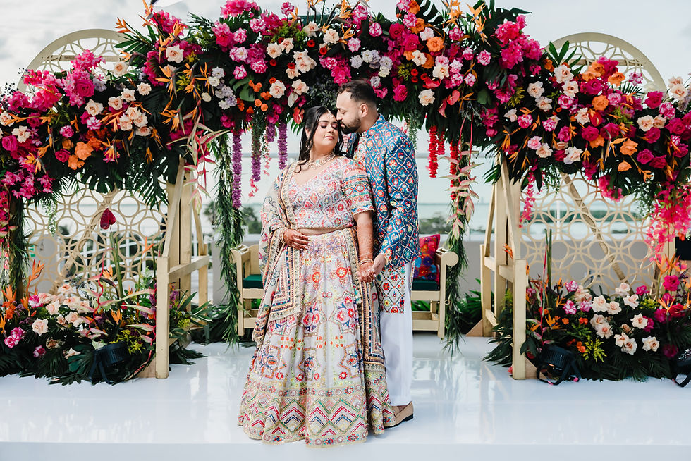 Indian couple posing beneath a vibrant floral arch during their destination wedding in Mexico, wearing colorful embroidered outfits with ocean views in the background.