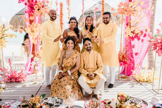 Bride and groom seated under a vibrant pink and orange Haldi mandap at Garza Blanca Cancun, surrounded by family members blessing them.