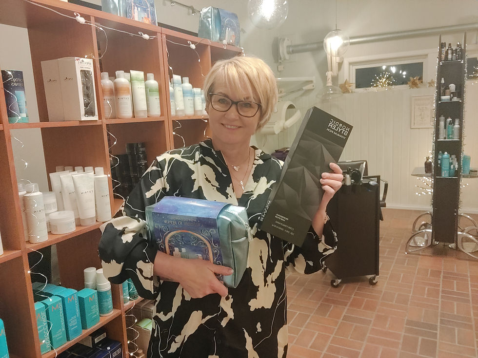 Dame med briller smiler og holder hårprodukter i hånda. Bak henne er frisersalong med produkter i hyllen.