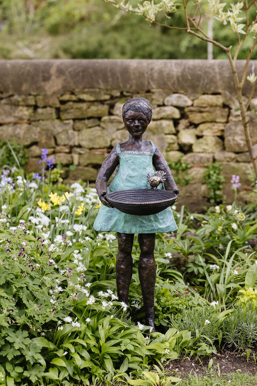 Thumbnail: Bronze girl in a green dress holding a birdbath bowl, standing amongst vibrant spring garden planting.