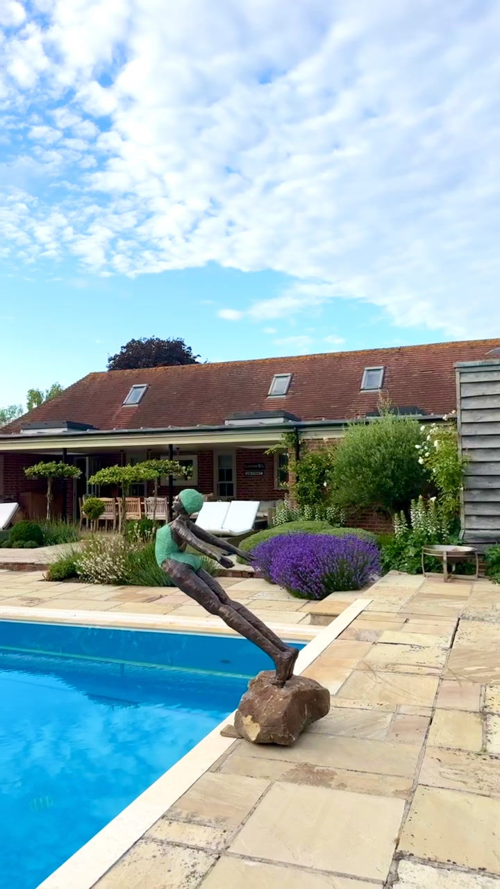 Thumbnail: Girl bronze sculpture leaning forward in a joyful dive pose, balanced on a rock beside a clear blue swimming pool.