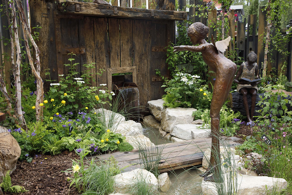 A striking bronze sculpture of a winged figure with arms outstretched, standing on a stone in a lush garden setting with a small stream, surrounded by greenery, moss-covered rocks, and tall trees under a clear blue sky. Ideal for garden art, outdoor sculptures, and nature-inspired decor enthusiasts.