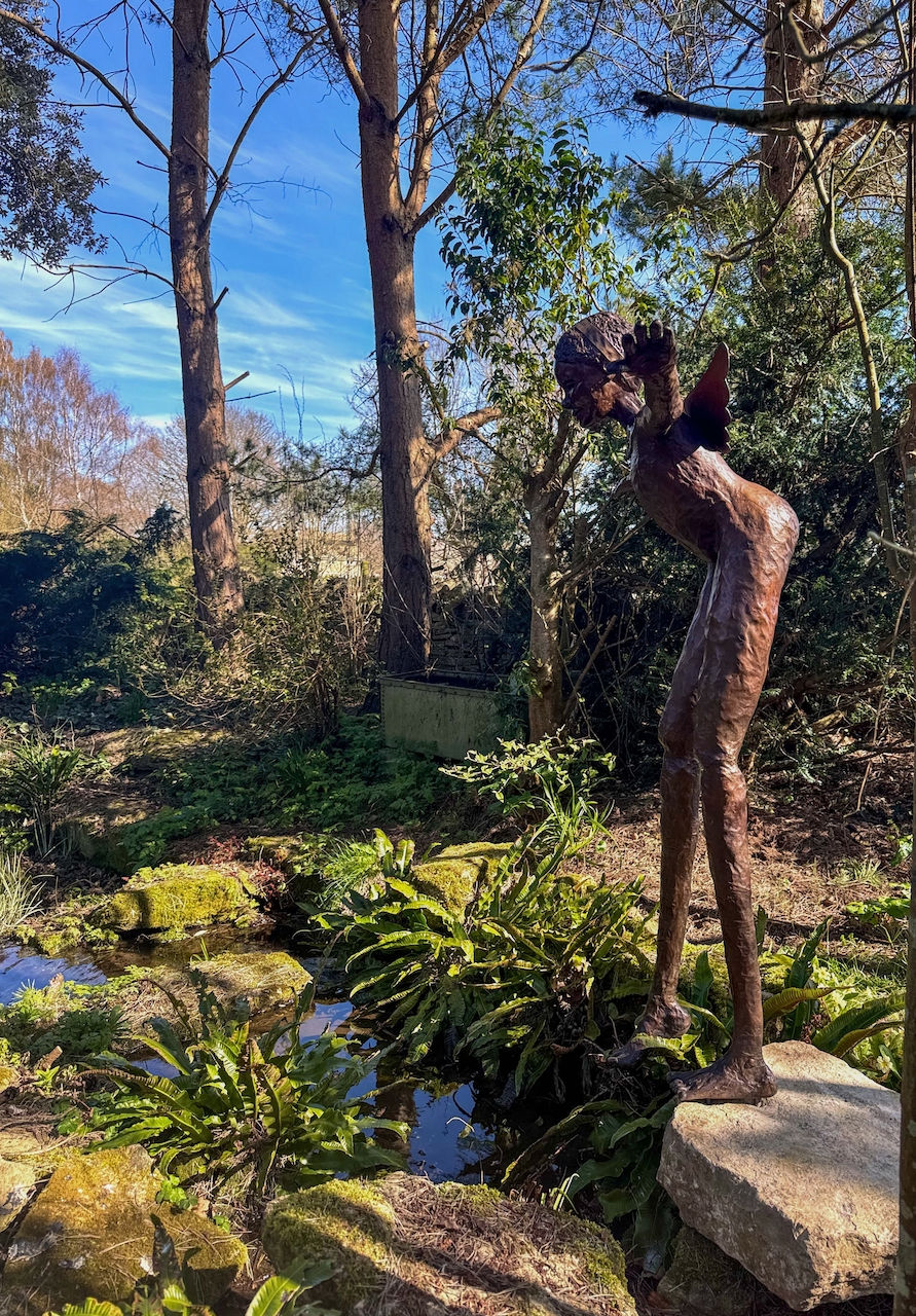 Thumbnail: Bare bronze feet of the winged girl poised on stone beside a garden stream