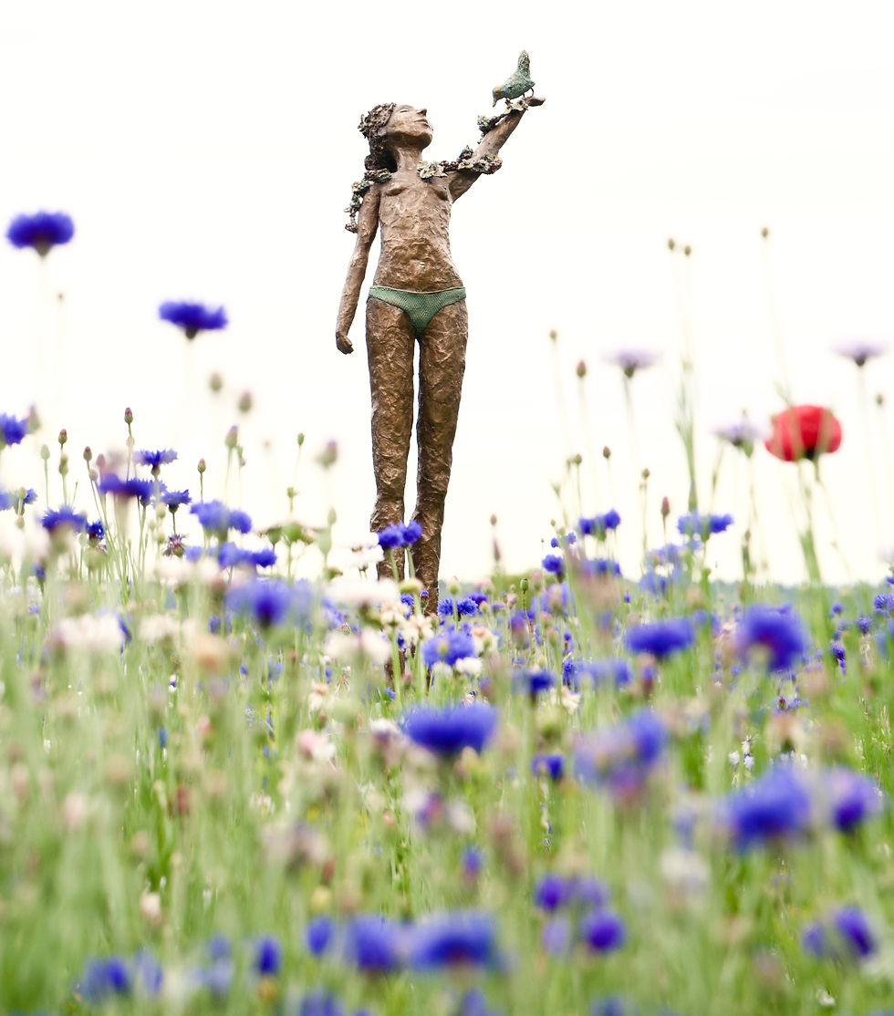 Bronze sculpture of a young girl standing in a wildflower meadow, raising her hand to a small bird perched on her fingers.