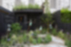 A vibrant garden display featuring bronze sculptures by Dawn Conn, including a central figure in a green swimsuit mid-leap, surrounded by smaller child sculptures with birds, set against a dark wooden backdrop with "DAWN CONN SCULPTURE" signage. Lush greenery, colorful flowers, and a young tree enhance the idyllic outdoor art scene. Perfect for garden sculpture, outdoor decor, and art lovers.
