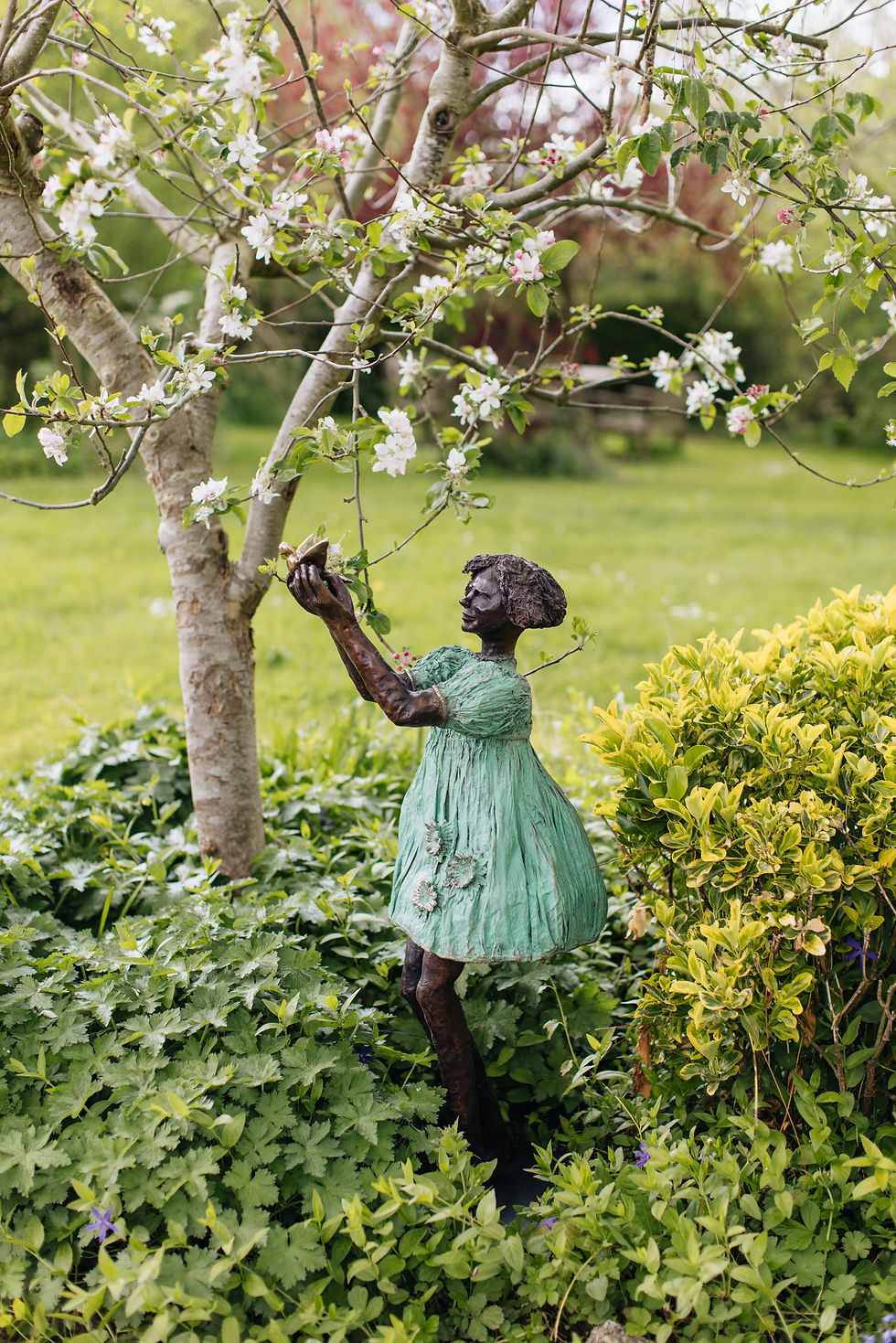 Thumbnail: Bronze sculpture of a girl holding a bird towards blooming apple branches, surrounded by vibrant garden greenery.