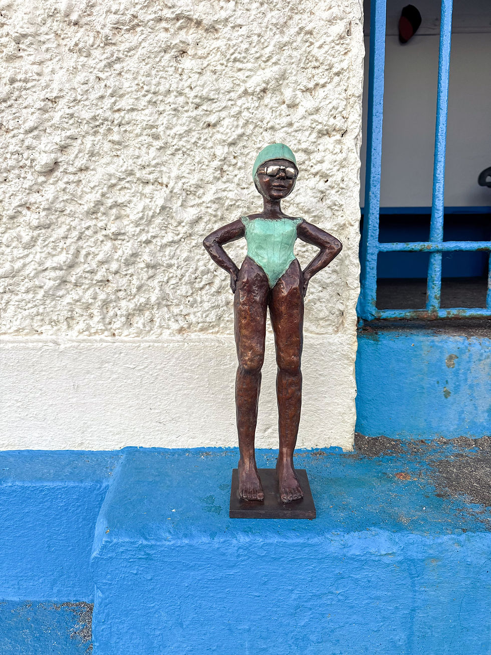 Thumbnail: “Bronze swimmer sculpture standing outside the Low Rock Swimmers changing hut with blue door; Dawn Conn coastal sculpture.
